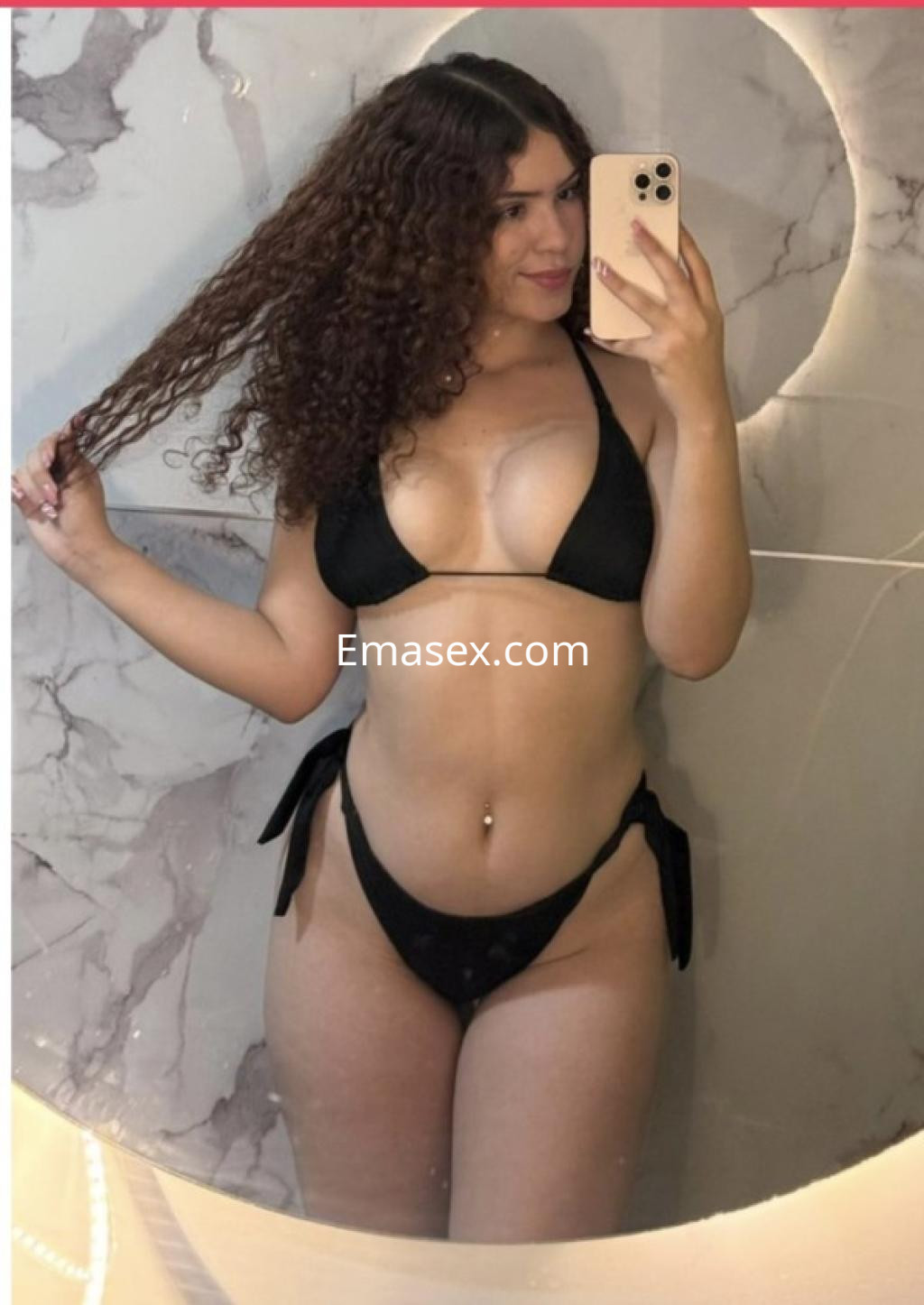 Escort de Pamplona posando frente a un espejo, con cabello rizado y un bikini negro que resalta su figura.