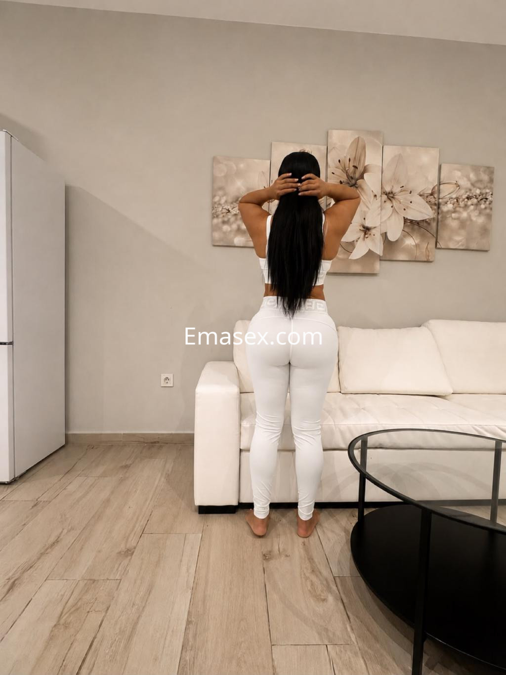 Escort de Córdoba con cabello largo y lacio, vestida con top blanco y pantalones ajustados, de pie en una sala moderna.