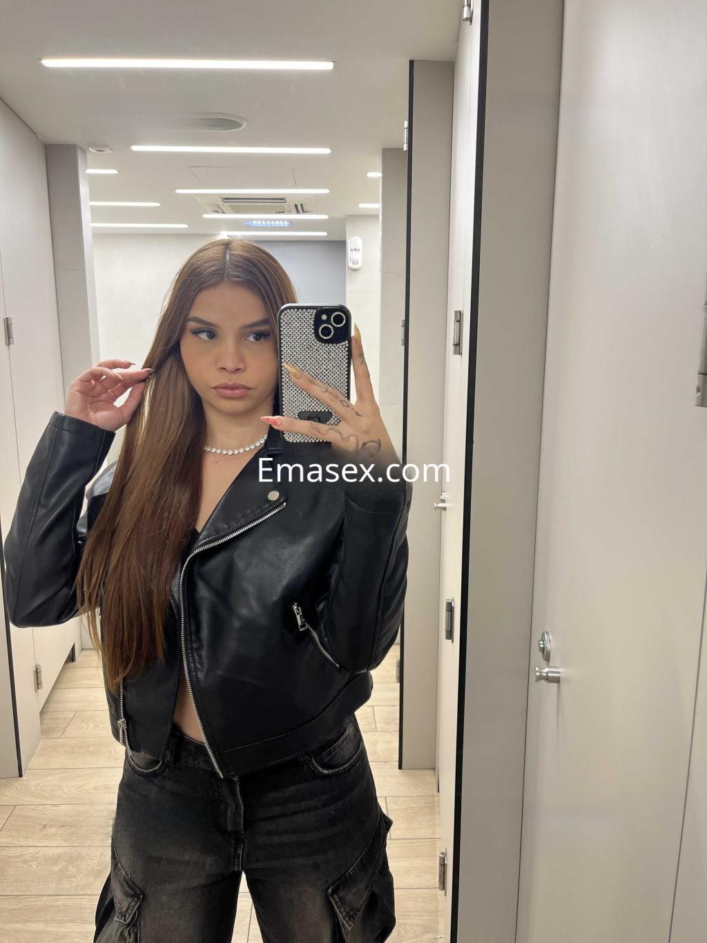 Escort en Almería posando frente al espejo con chaqueta de cuero negra y pantalones oscuros, luciendo cabello largo.