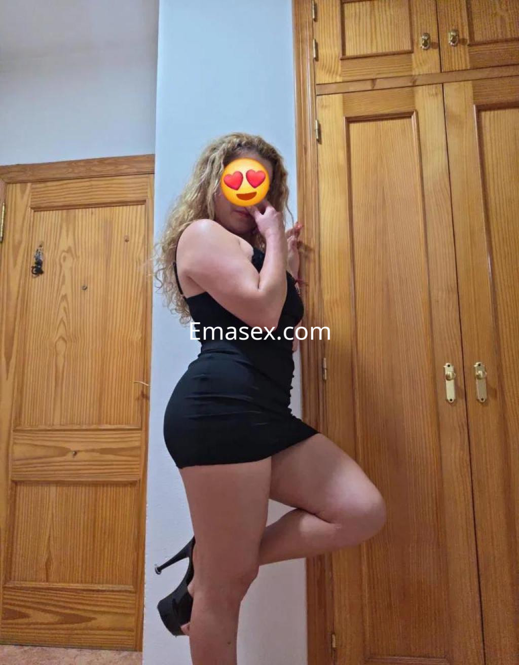 Escort en Almería luciendo un vestido negro ceñido y tacones altos, posando con una mano en la cara.