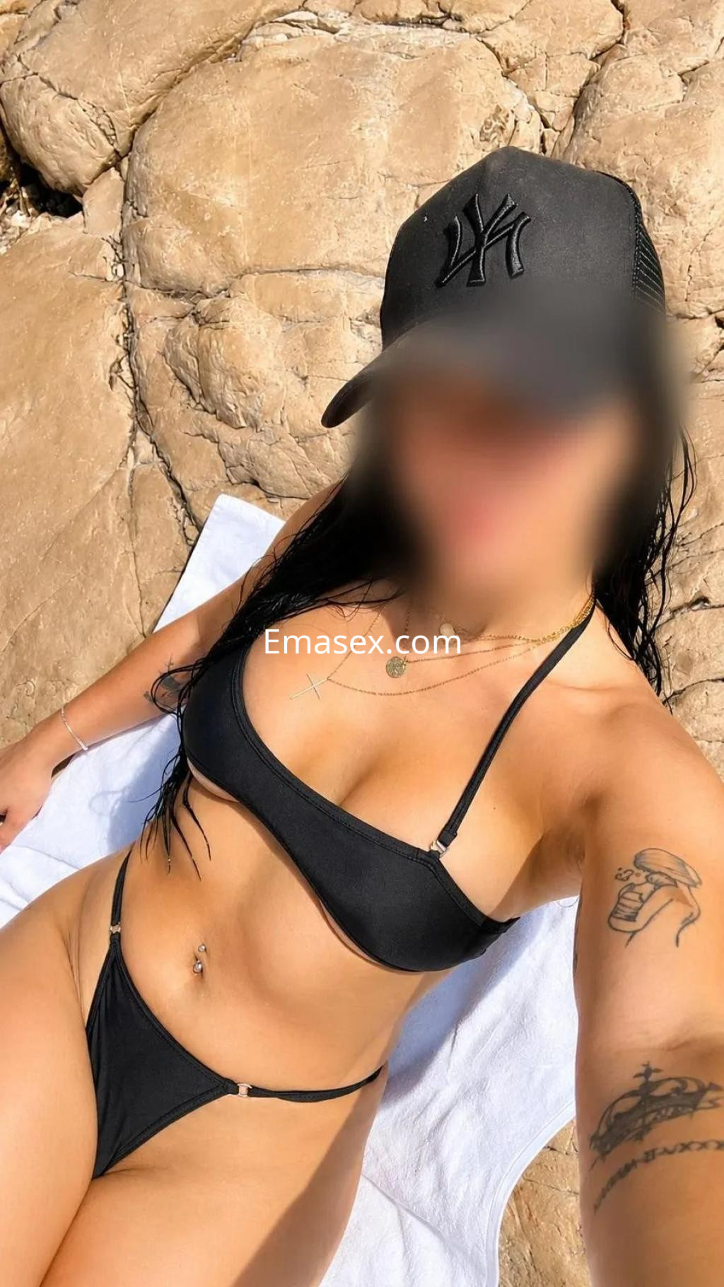 Escort en Murcia tomando el sol en la playa, luciendo un bikini negro y una gorra, con un fondo rocoso.