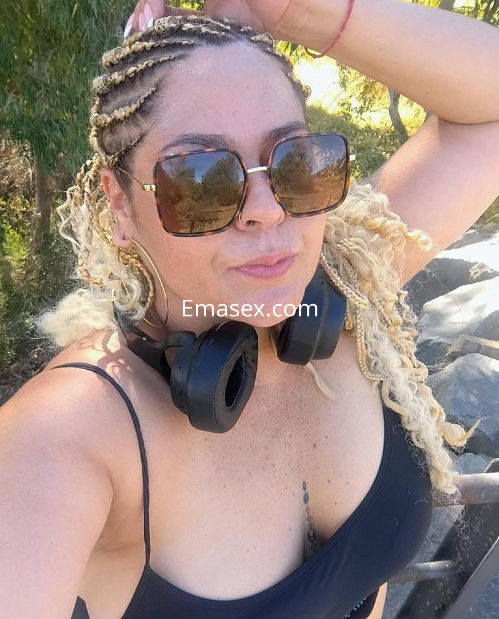 La puta luce un top negro en Oviedo, con trenzas rubias y gafas de sol grandes, en un entorno al aire libre.
