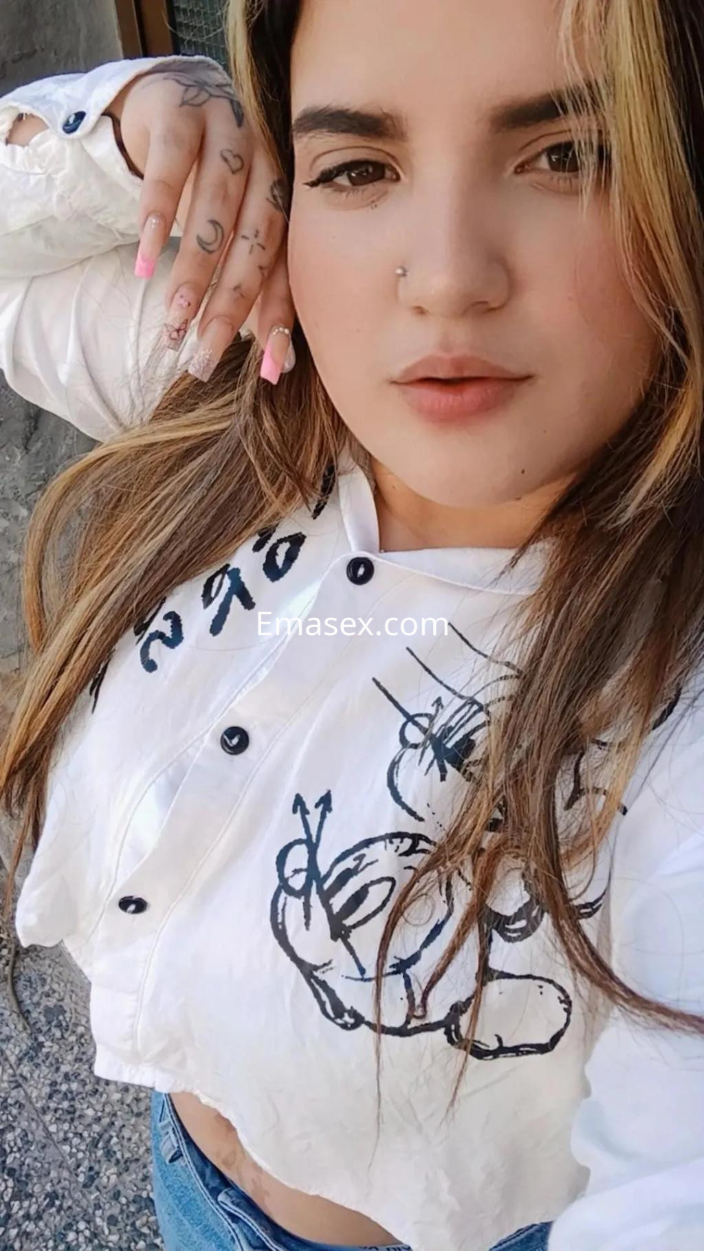 La puta en Zaragoza posa con una blusa blanca y jeans, mostrando uñas largas y un maquillaje natural.