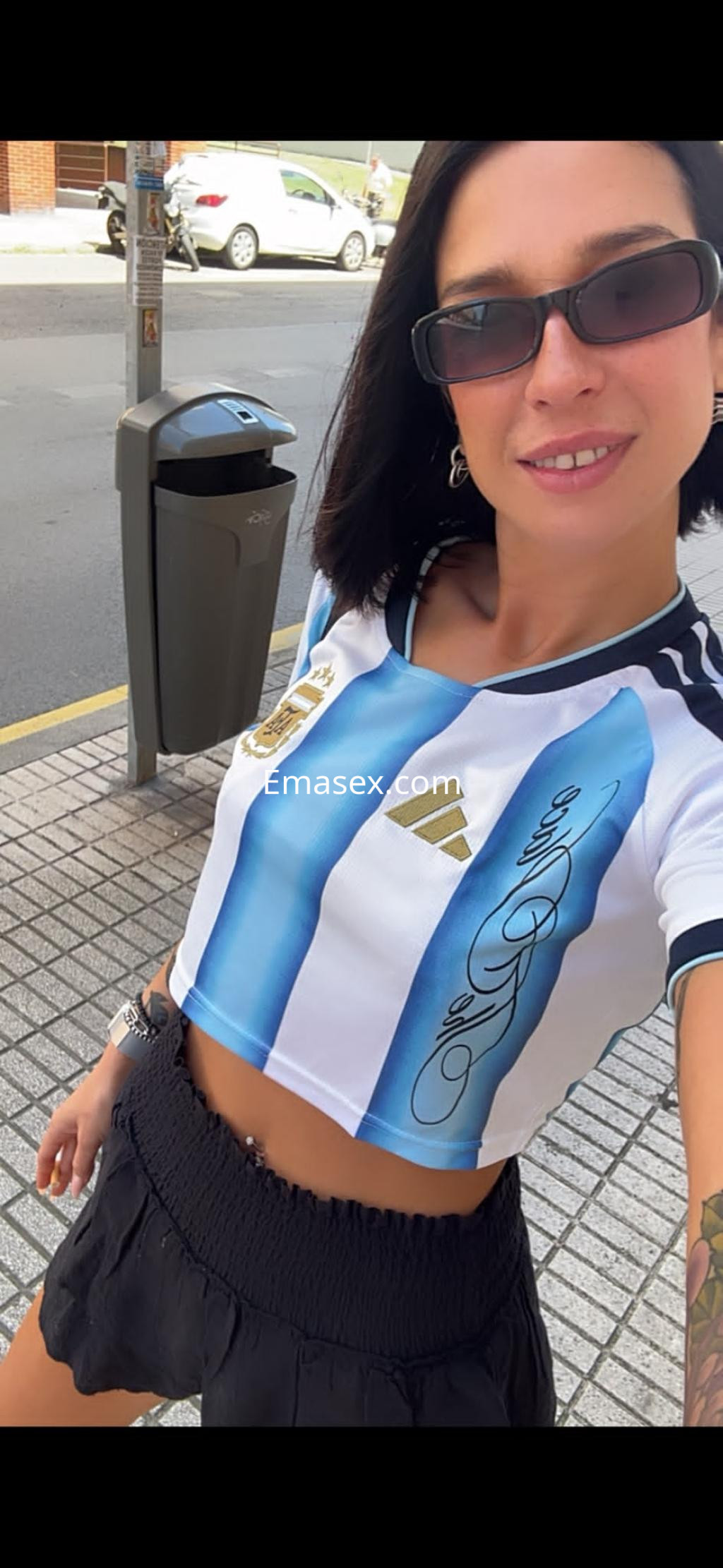 Puta en Santander luce un crop top blanco con rayas azules y negras, y una falda corta negra, sonriendo a la cámara.