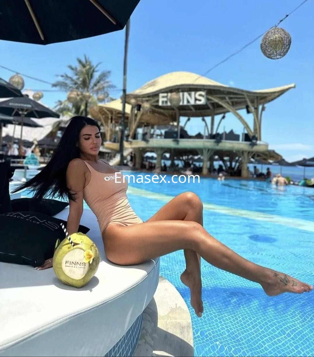 Puta con traje de baño claro, sentada junto a una piscina en Alicante, disfrutando del sol y un ambiente tropical.