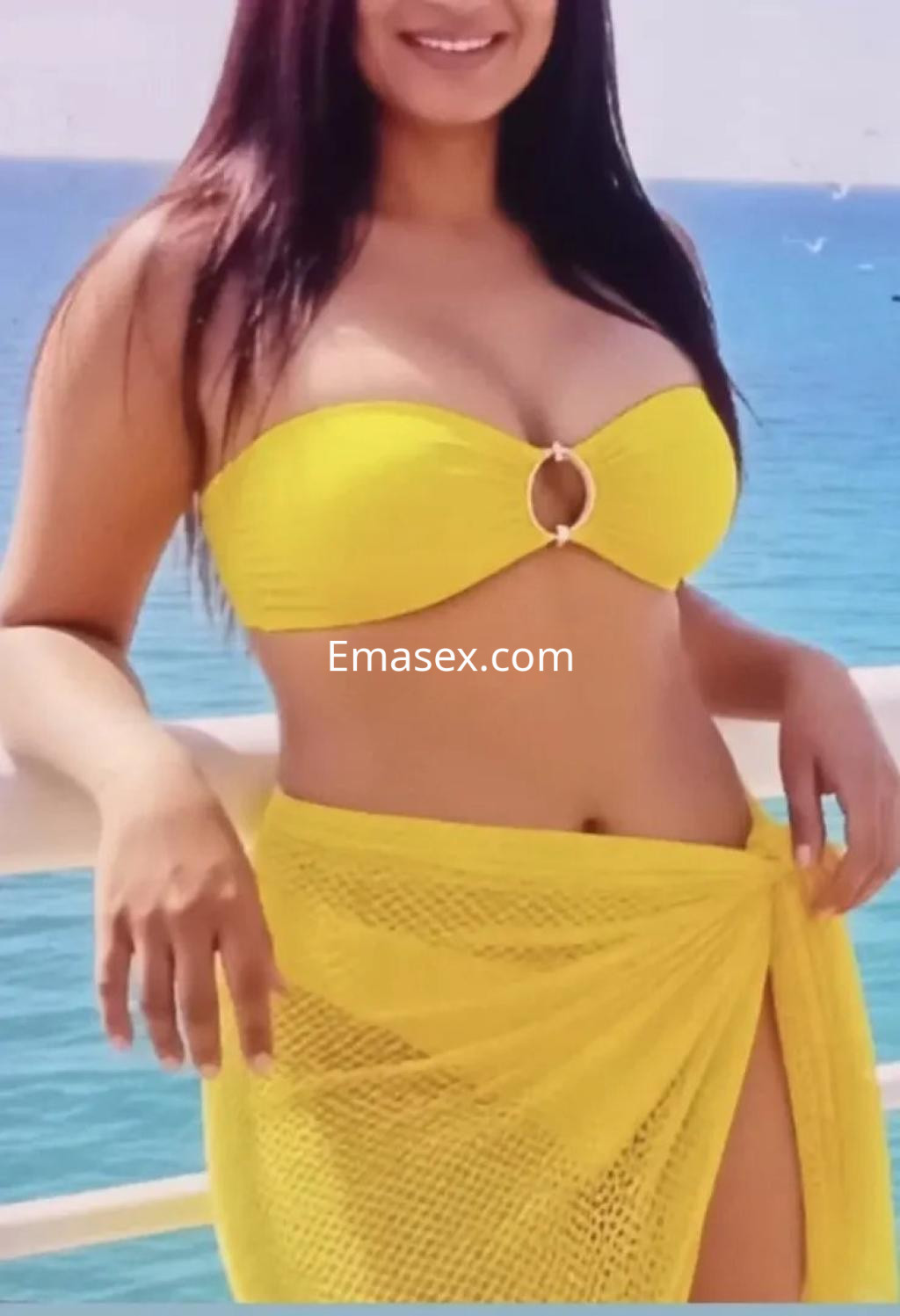 Escort con cabello largo y oscuro, vistiendo un bikini amarillo, posando frente al mar en Málaga.