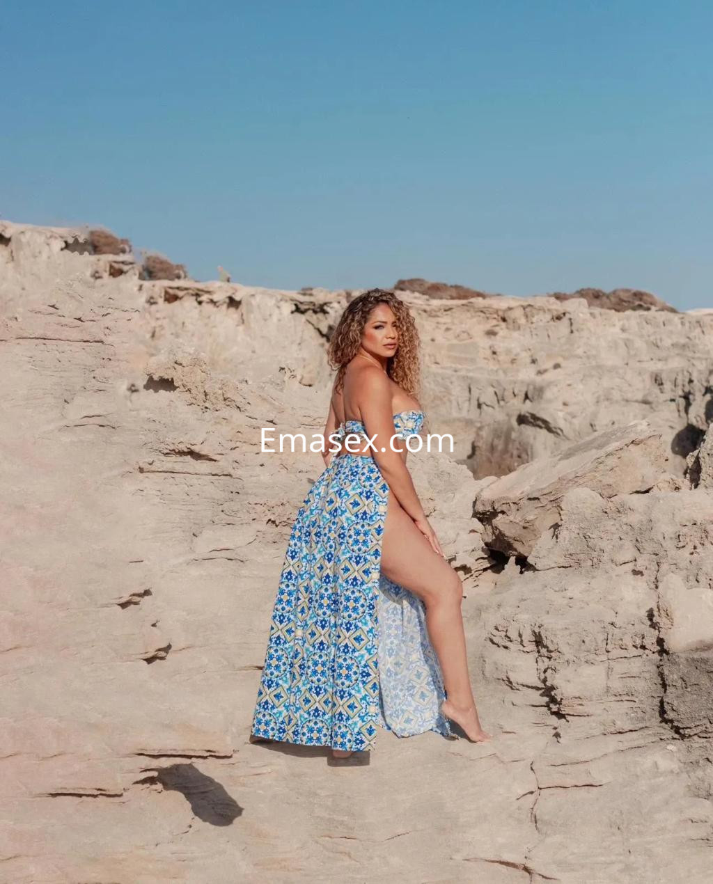 Escort en Valencia con cabello rizado, vistiendo un vestido largo azul y blanco, posando en una playa rocosa bajo el sol.