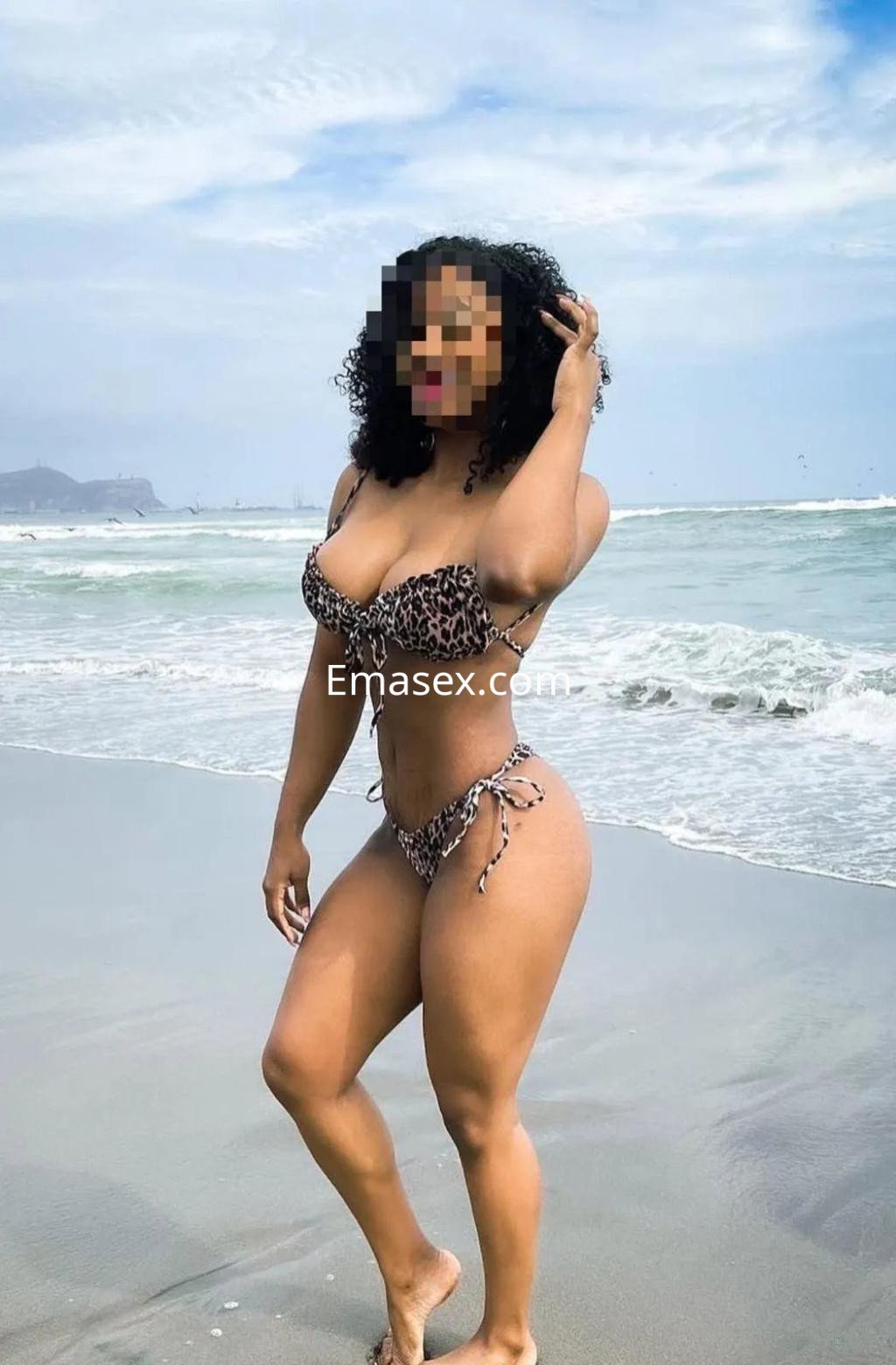 Escort en Valencia con bikini de estampado animal, posando en la playa con el mar de fondo y cabello rizado.
