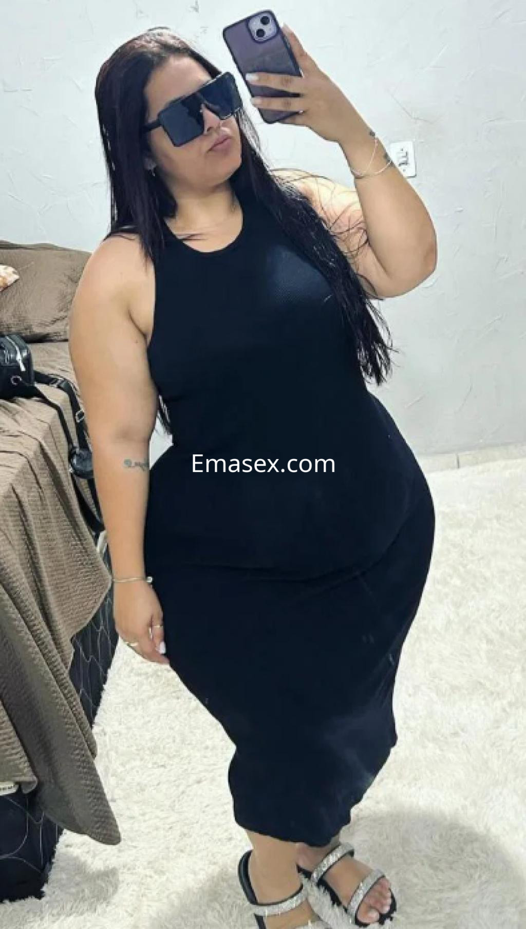 Escort en Logroño, posando con un vestido negro ajustado y gafas de sol, en un entorno interior.