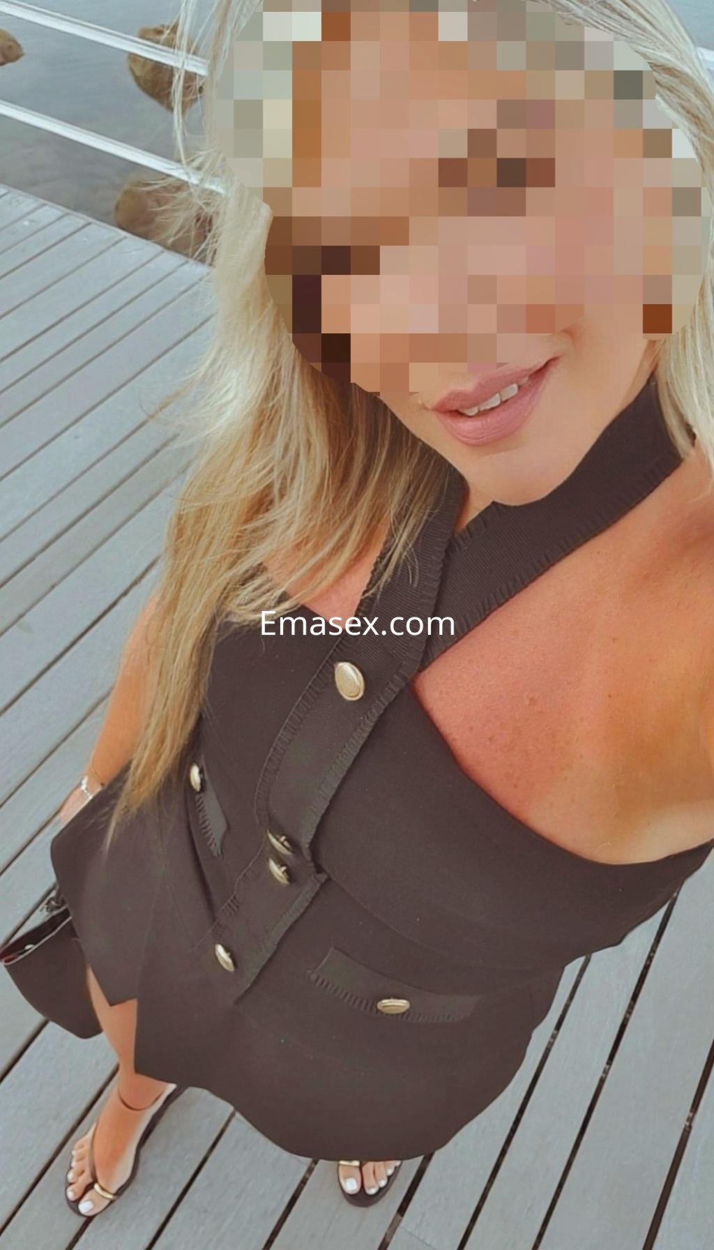 Escort en Coruña, con cabello rubio y lacio, viste un vestido negro sin mangas, posando en una terraza de madera.