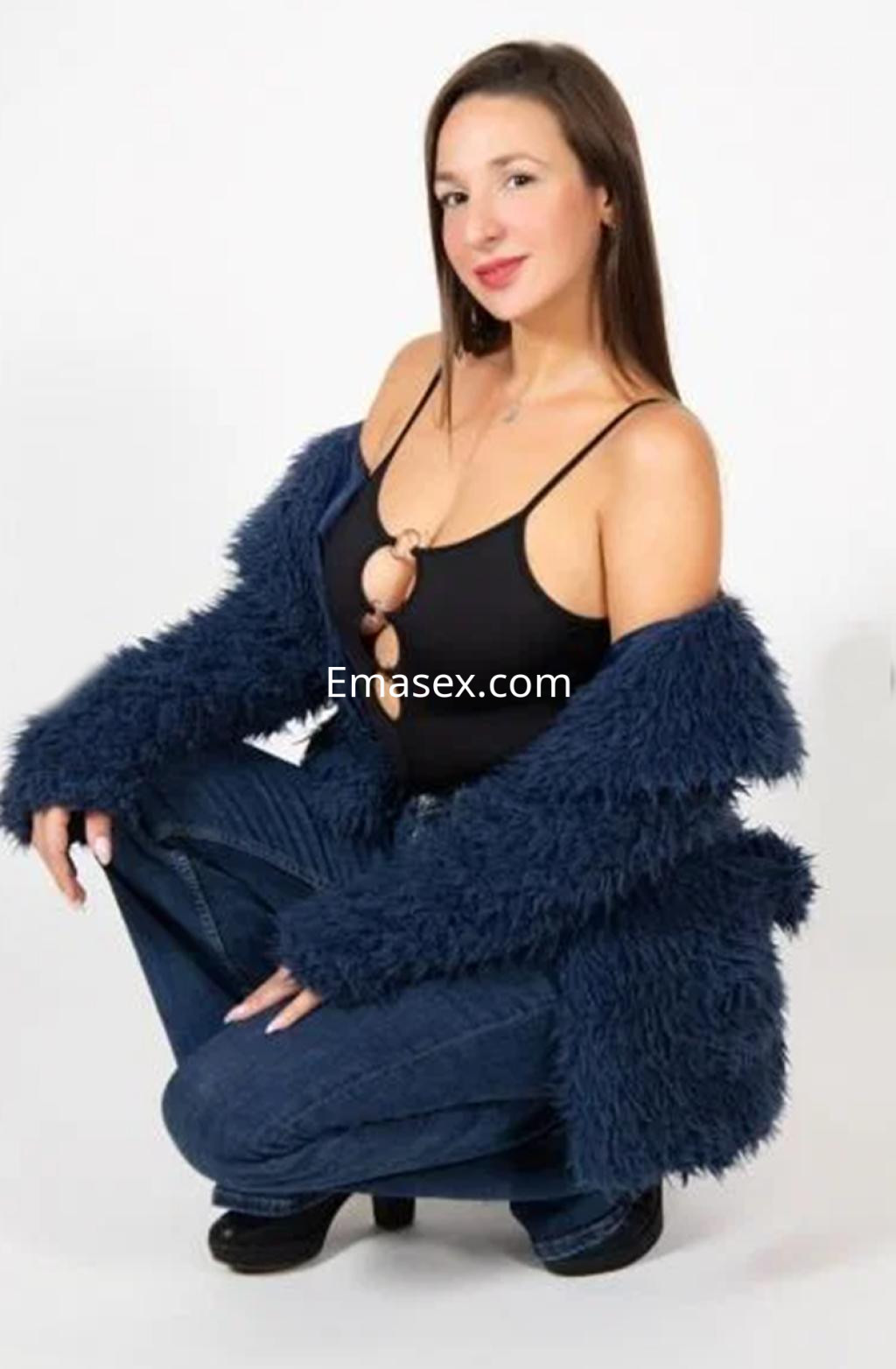 Escort en Santander, posando en cuclillas con un top negro y chaqueta de peluche azul, luciendo un estilo moderno.