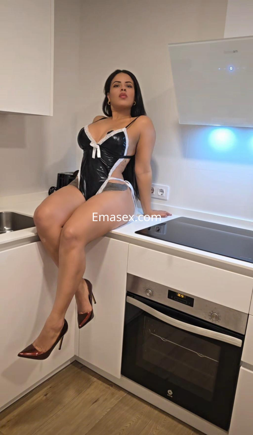 La puta posando en una cocina moderna, viste un conjunto de látex negro con detalles blancos y tacones rojos.