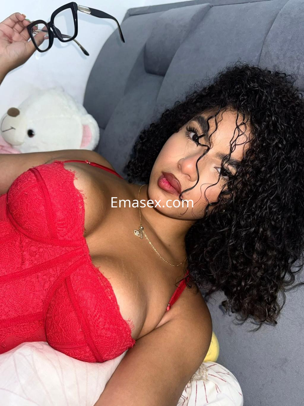 Escort en Vigo posando sobre la cama con un conjunto de lencería roja, sosteniendo unas gafas y con rizos oscuros.