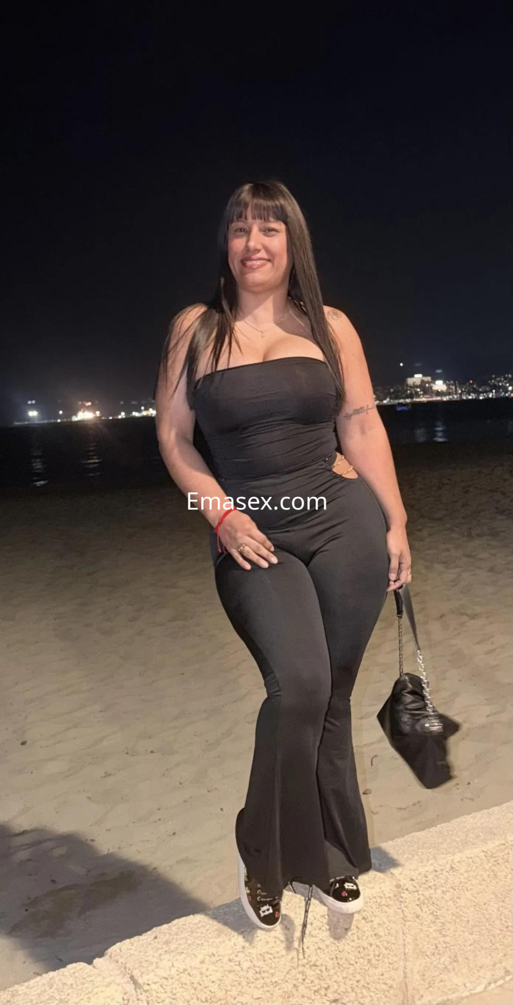 Puta de cabello largo y lacio, viste un ajustado mono negro, posando junto a la playa en Palma de Mallorca.