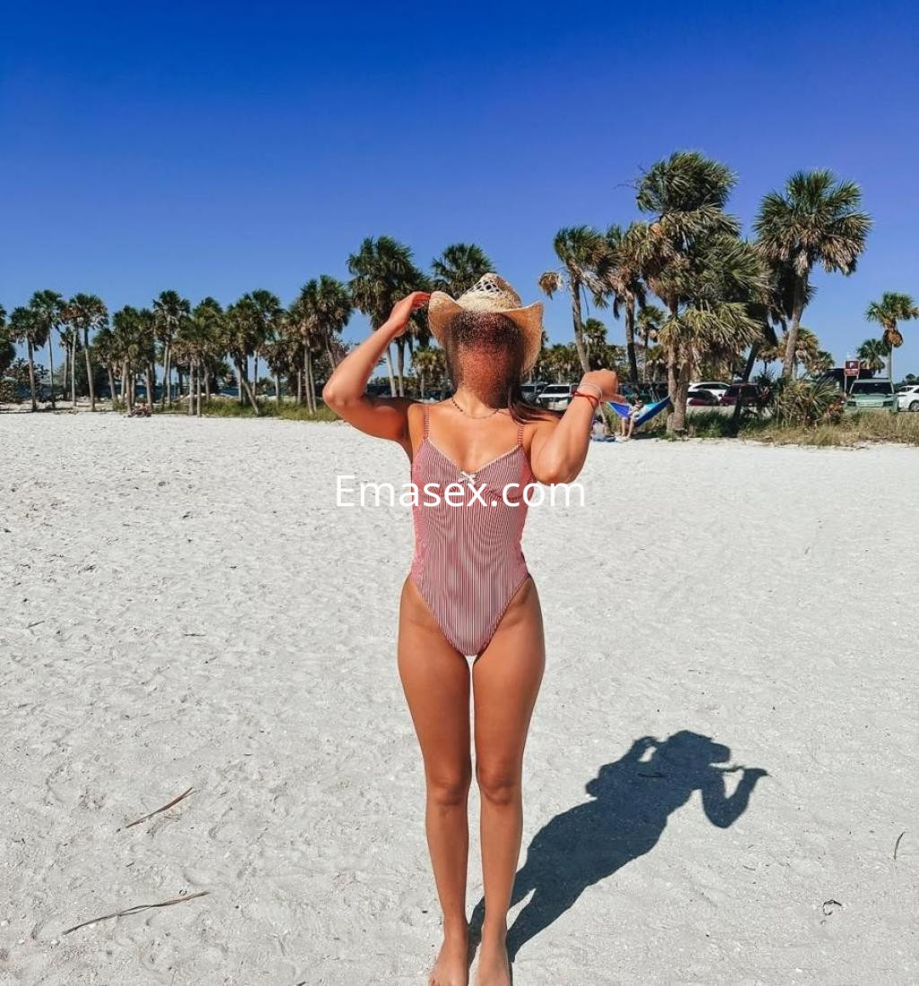 Escort en Coruña, de espaldas en la playa, lleva un bañador de rayas y un sombrero, con palmeras al fondo.
