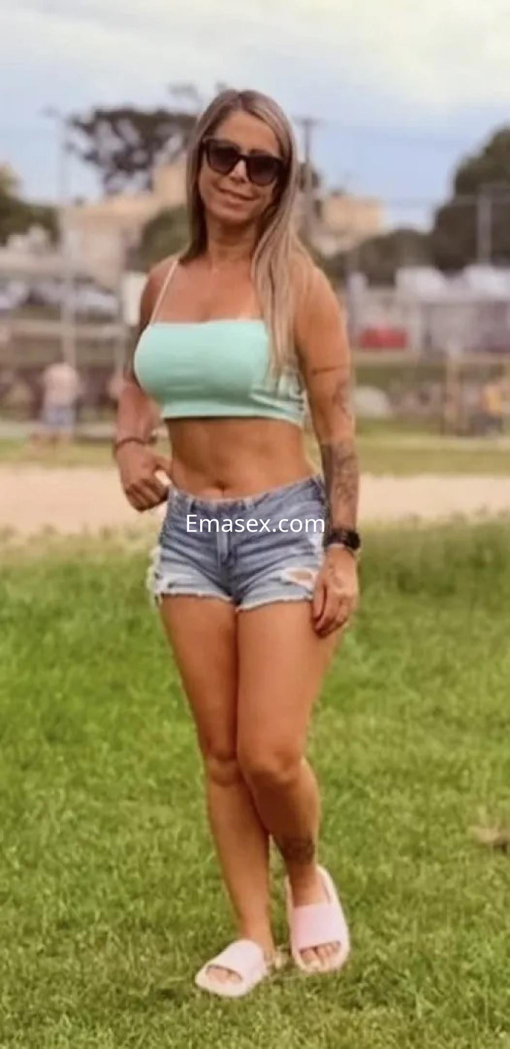 Escort en Coruña con cabello largo y rubio, luciendo un top verde y shorts de mezclilla, de pie en un parque.