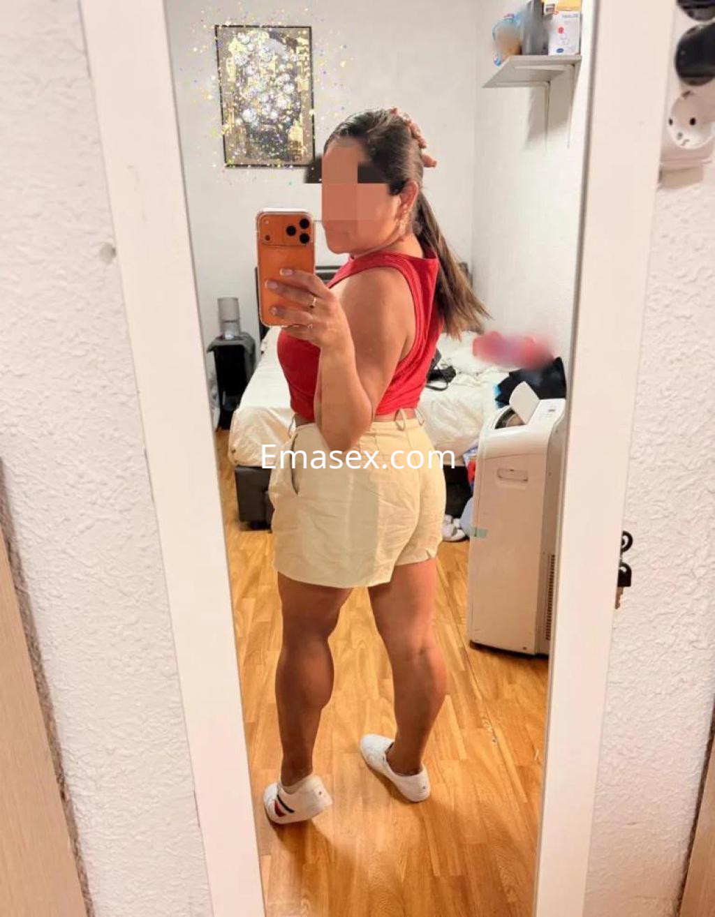 Escort en Lleida posando frente a un espejo, viste una blusa roja y pantalones cortos claros, con cabello amarrado.