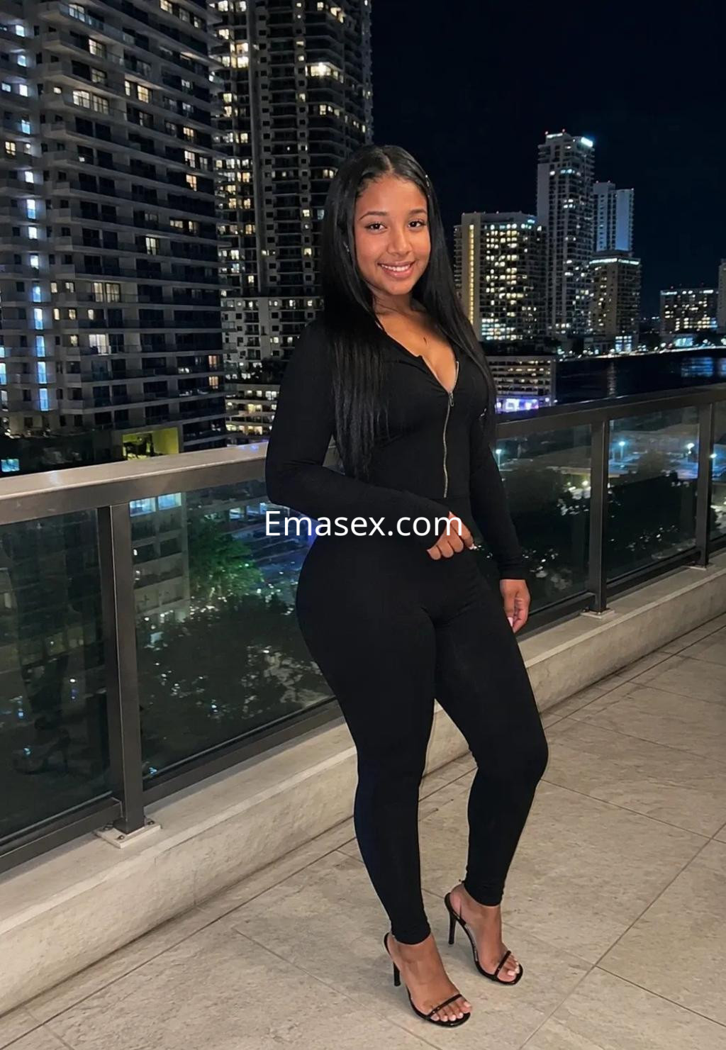 Escort en Zaragoza luciendo un ajustado conjunto negro y tacones, posando con un fondo urbano iluminado.