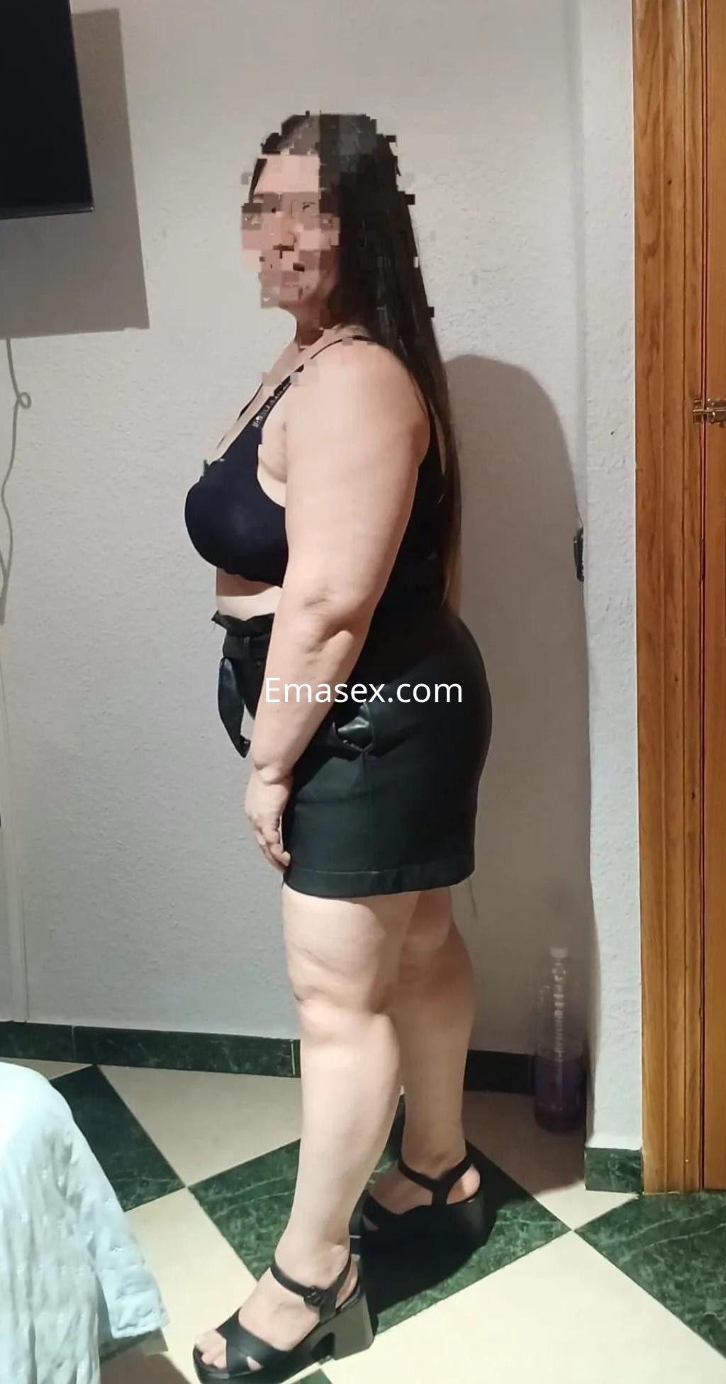 Puta de Albacete con cabello largo, viste un top negro y una minifalda de cuero, posando de perfil.