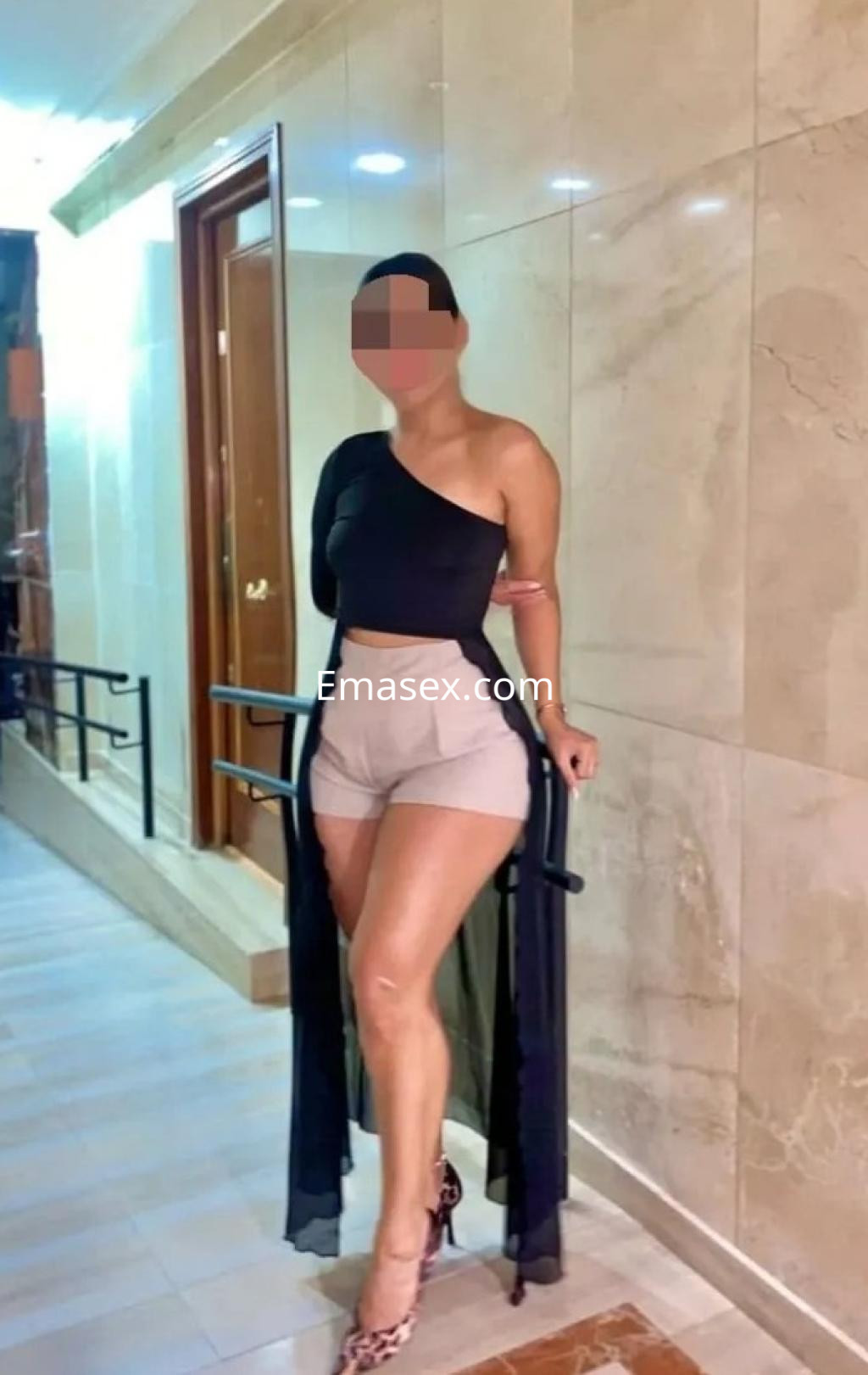 Escort en Albacete con blusa negra de un solo hombro y shorts claros, posando en un pasillo elegante.