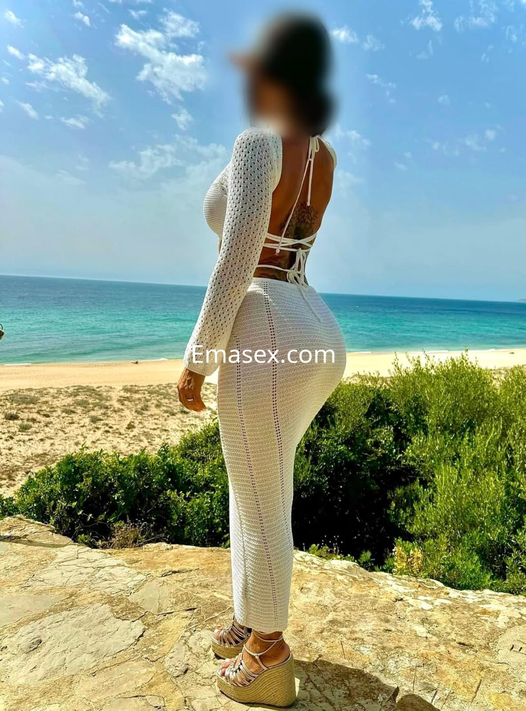 Escort de Bilbao posando de espaldas en la playa, con un traje blanco de punto y sandalias altas.