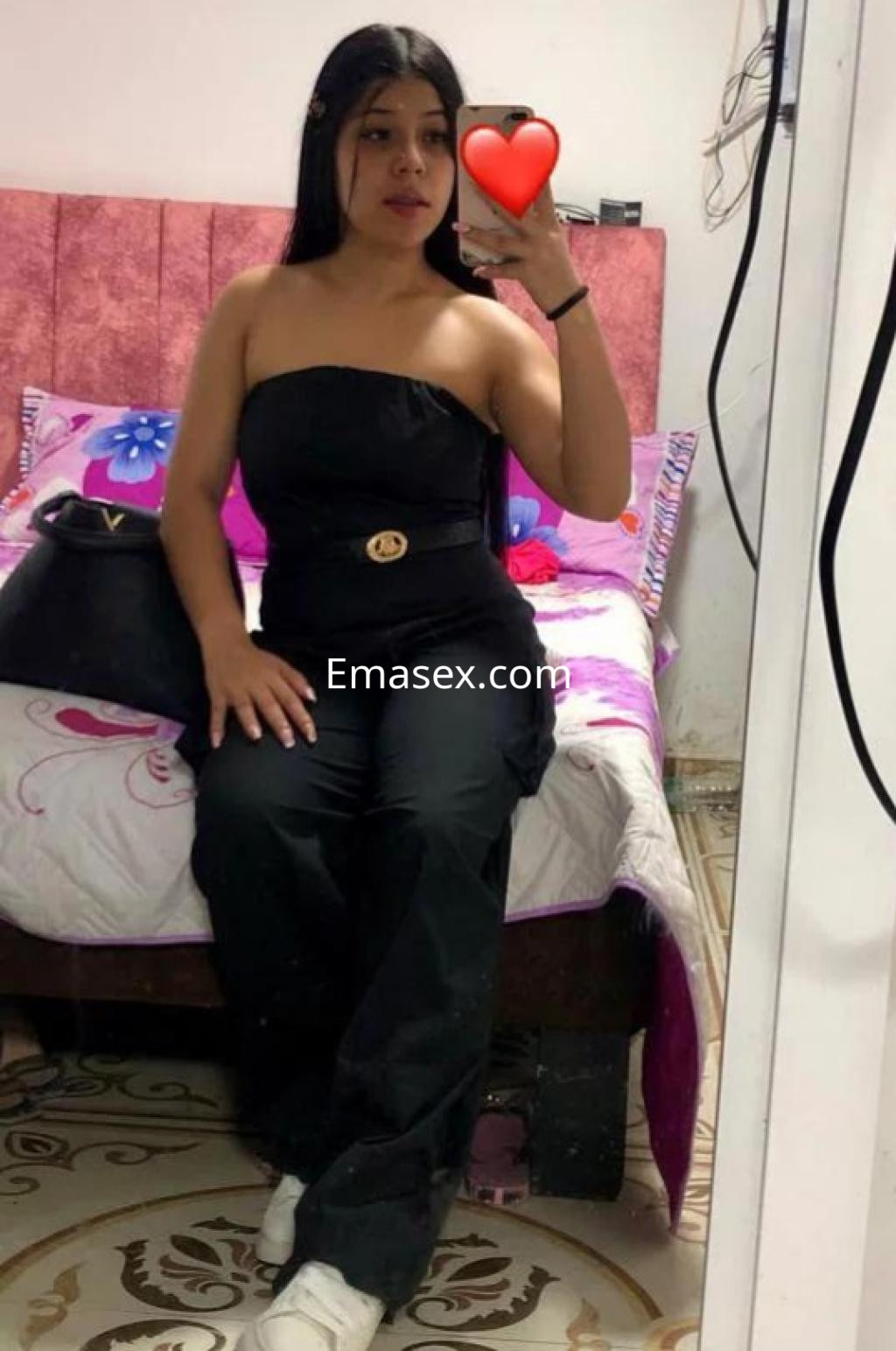 Escort en Castellón viste un conjunto negro sin mangas, posando frente a un espejo con una cama de fondo.