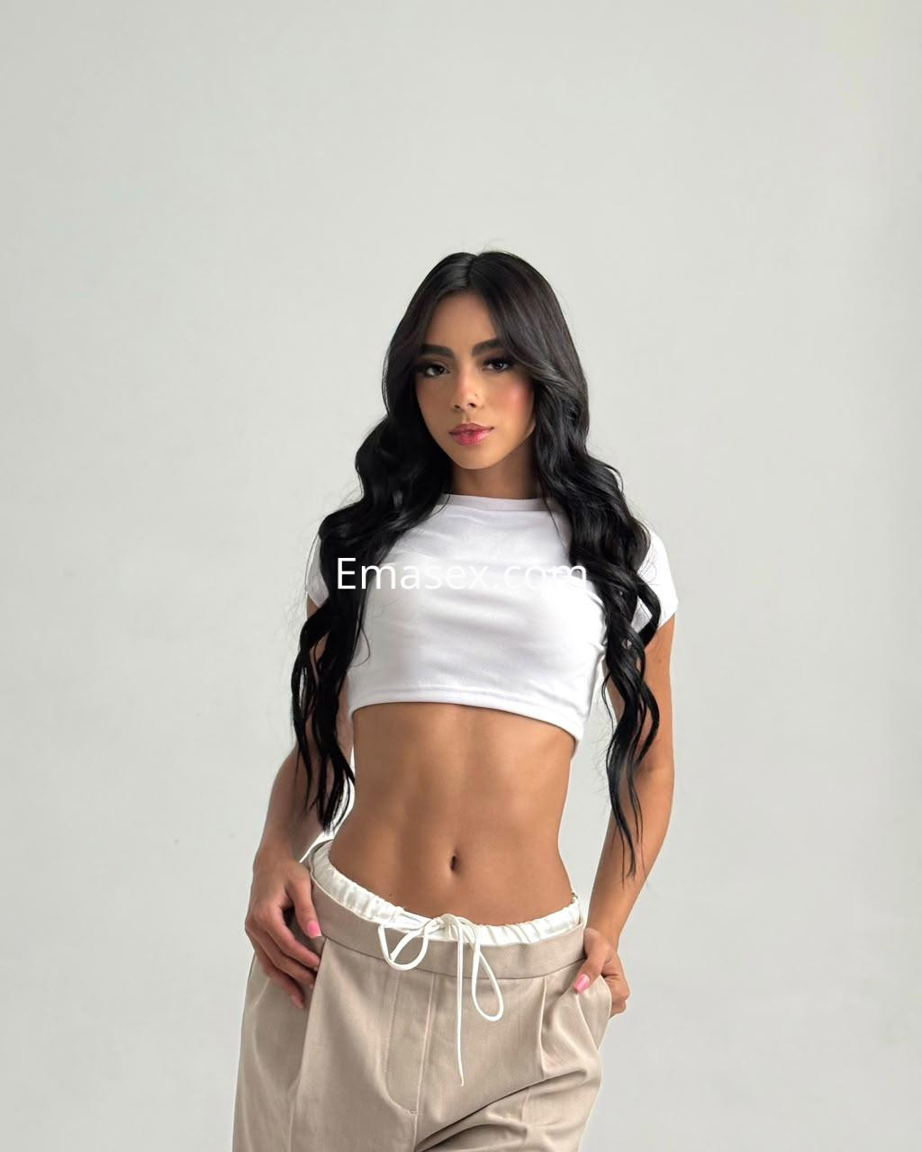 La puta viste una camiseta blanca y pantalones sueltos, posando con confianza en un fondo neutro en Palma de Mallorca.