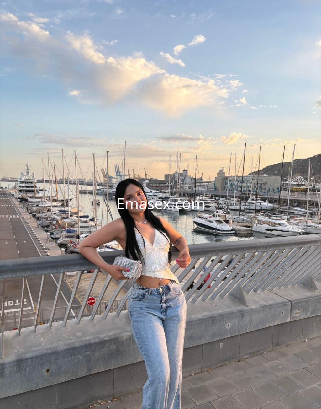 Escort en Girona posando junto al puerto al atardecer, con blusa brillante y jeans ajustados.