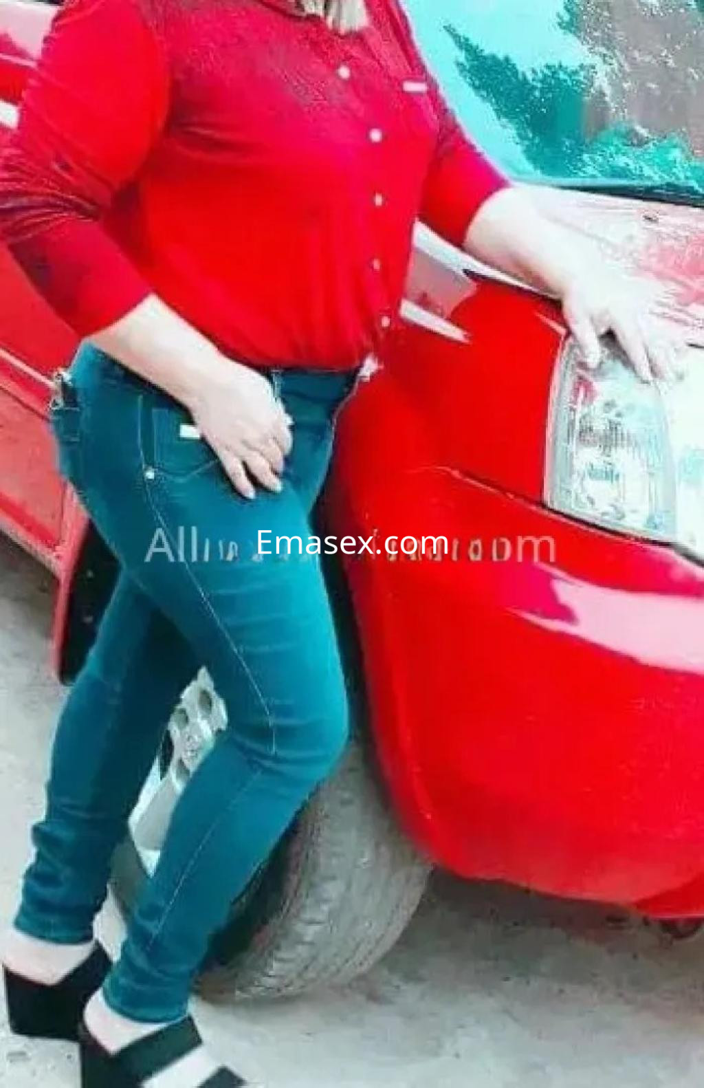 Escort en Albacete posando junto a un coche rojo, viste una blusa roja y jeans ajustados.