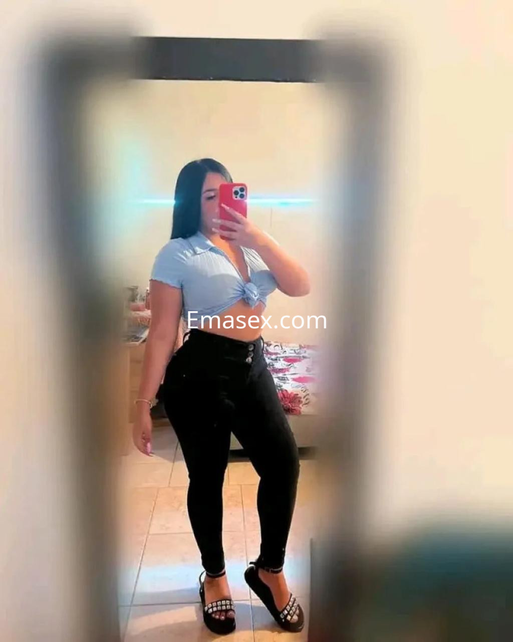 La puta en Alicante posa frente al espejo con blusa corta azul y pantalones ajustados negros.