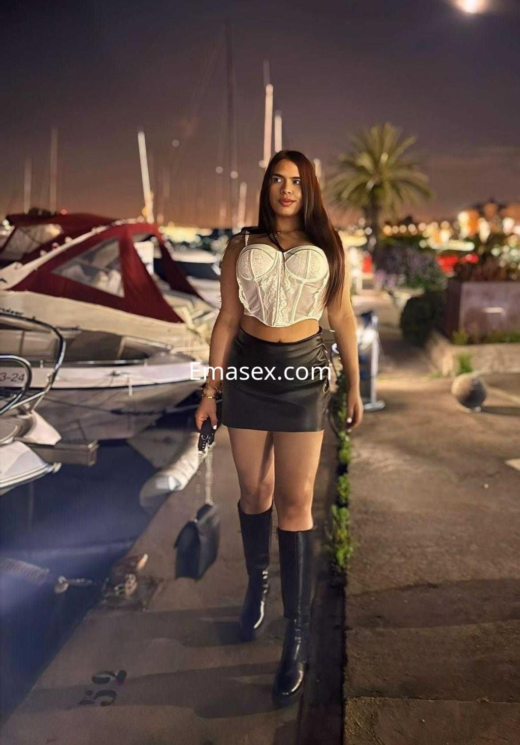 Escort caminando junto a un puerto en Barcelona, vistiendo un top blanco, falda corta y botas altas.