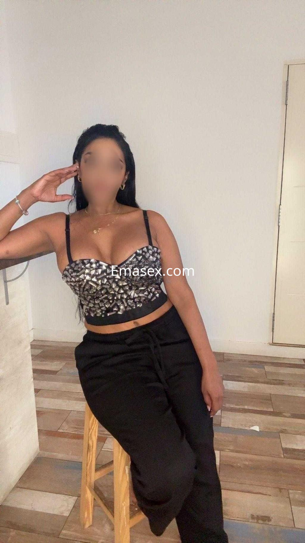 Escort en Burgos, sentada sobre un taburete, viste un top de tirantes y pantalones negros, con pelo oscuro.