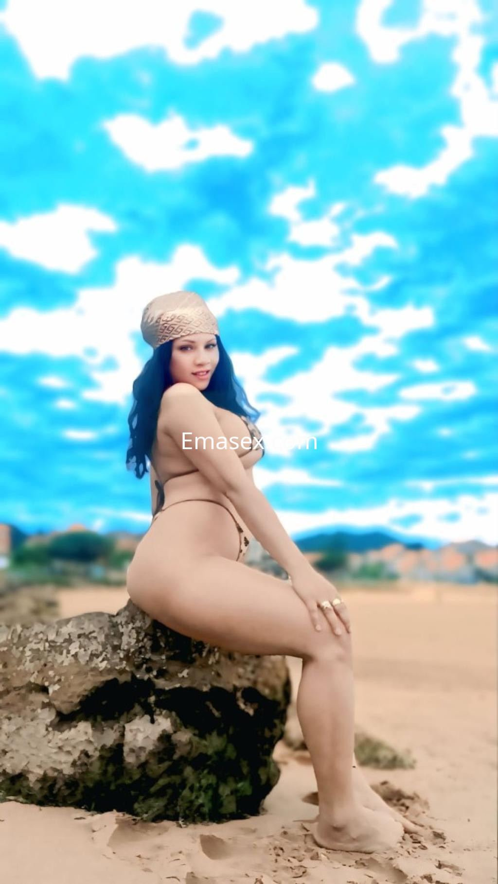 Escort en Burgos, posando de perfil sobre una roca en la playa, con un bañador negro y un pañuelo en la cabeza.