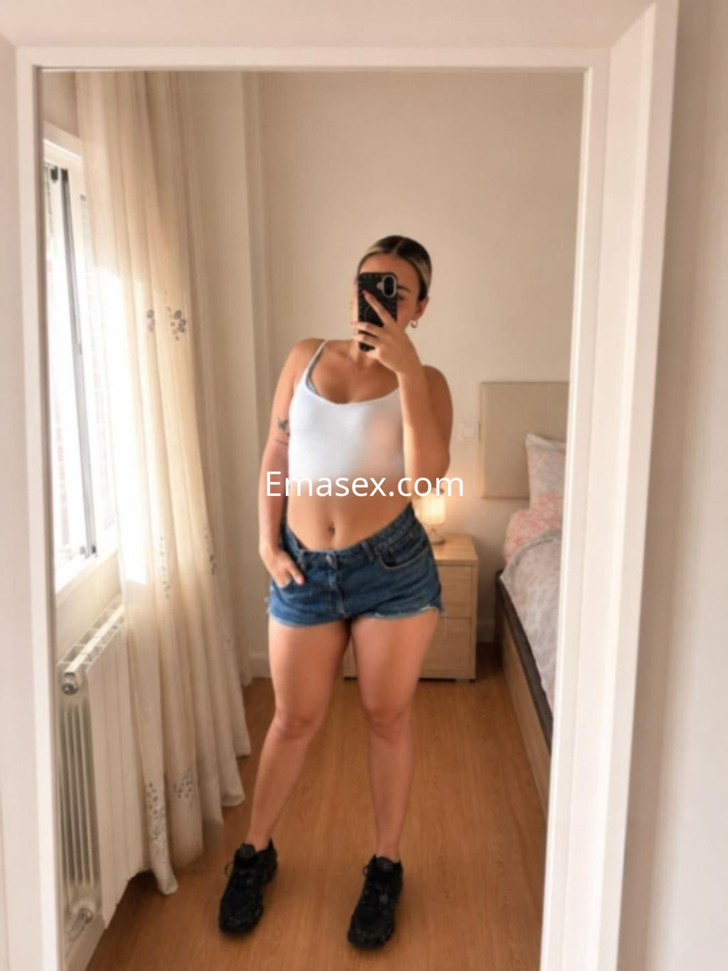 Escort en Burgos, posando frente al espejo con una camiseta sin mangas y shorts de mezclilla, mostrando una mirada confiada.