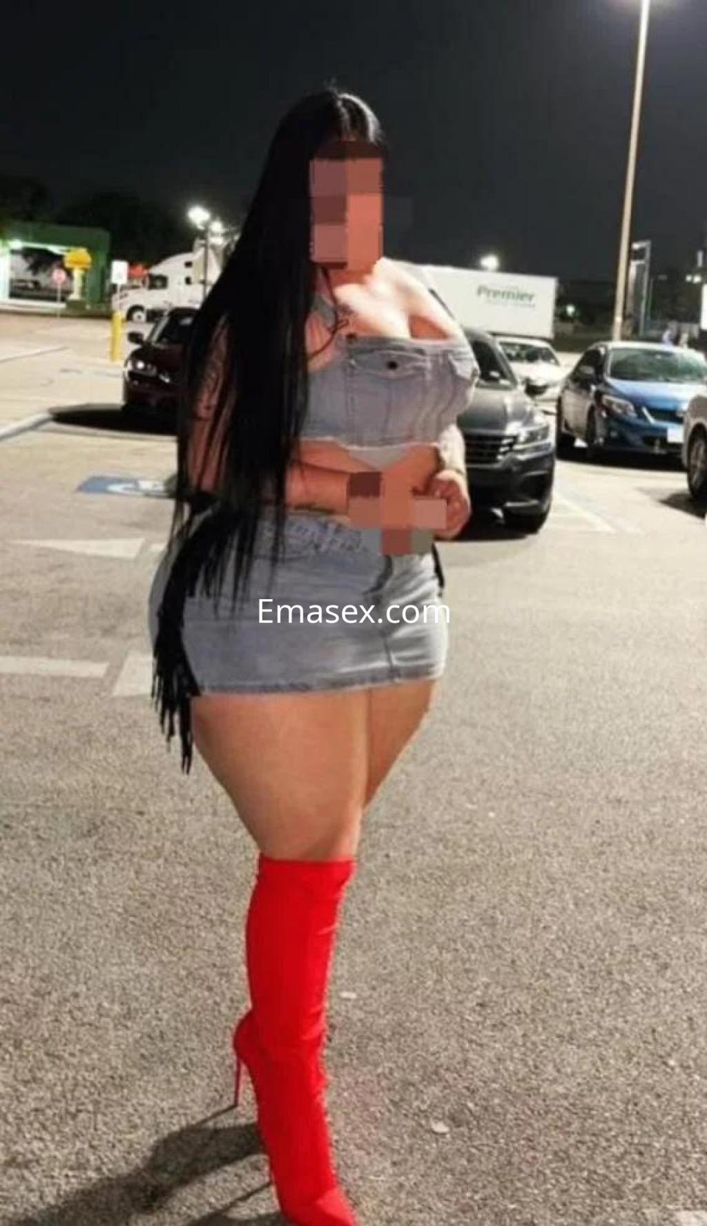 Escort en Cádiz, luciendo un vestido corto de mezclilla y botas rojas altas, posando en un área de estacionamiento.
