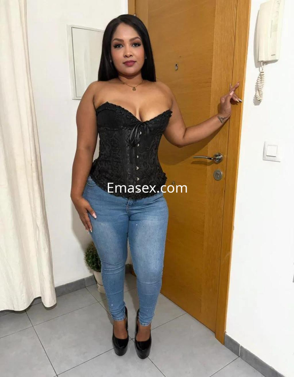 Escort en Cádiz, posando junto a una puerta con un corset negro, jeans ajustados y tacones altos.