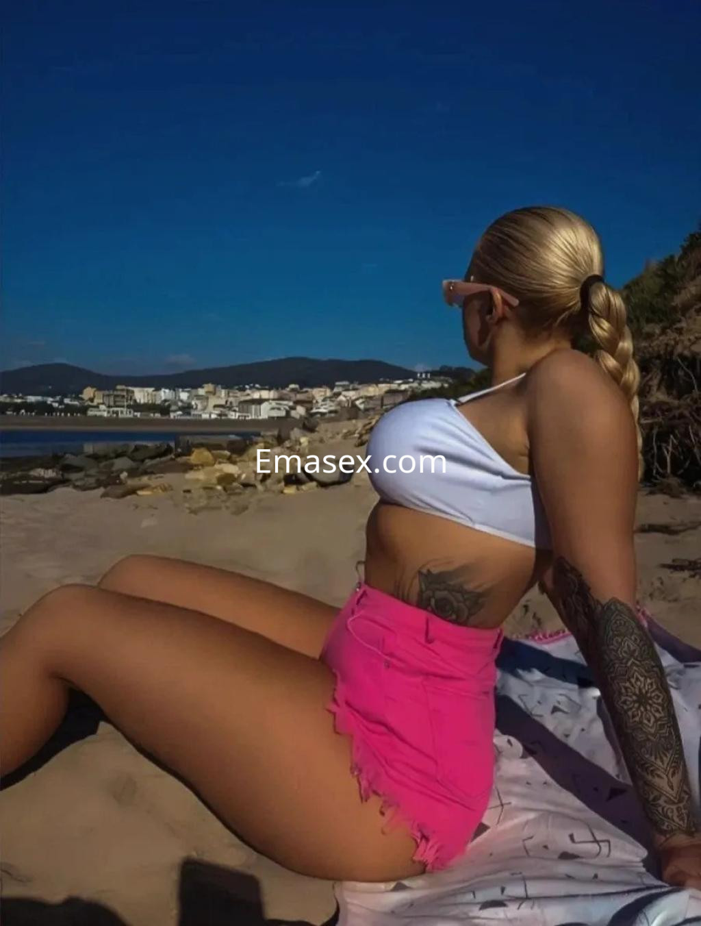 Escort en Coruña, sentada en la playa con un top blanco y una falda rosa, mirando hacia el mar.