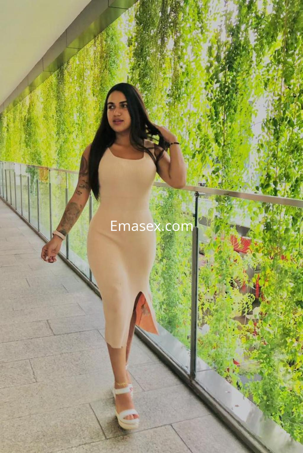 Escort en Girona con vestido ajustado de color nude y sandalias blancas, caminando entre vegetación.