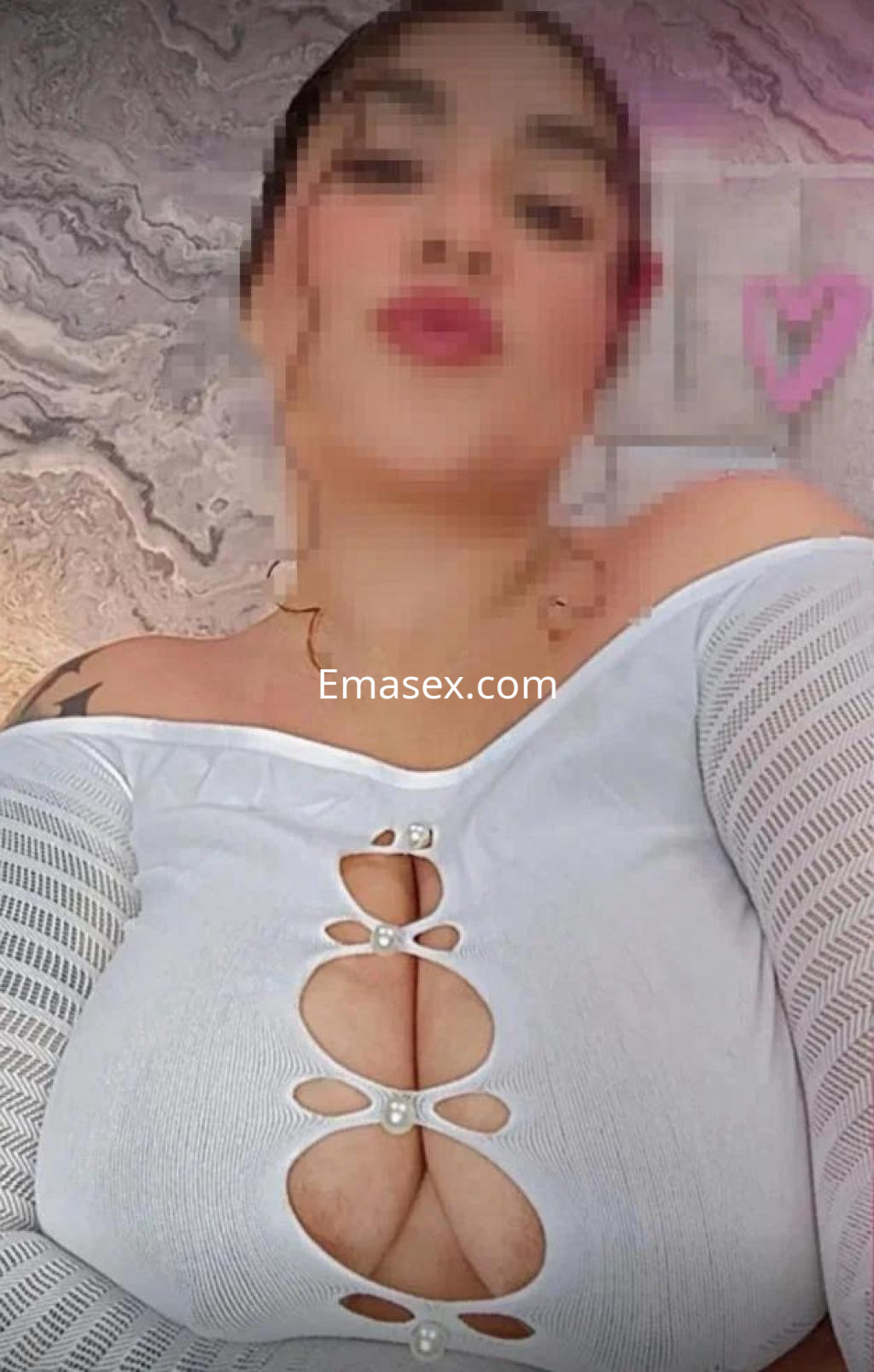 Escort en Girona viste una blusa blanca con detalles en perlas, posando de manera sugestiva ante un fondo decorativo.