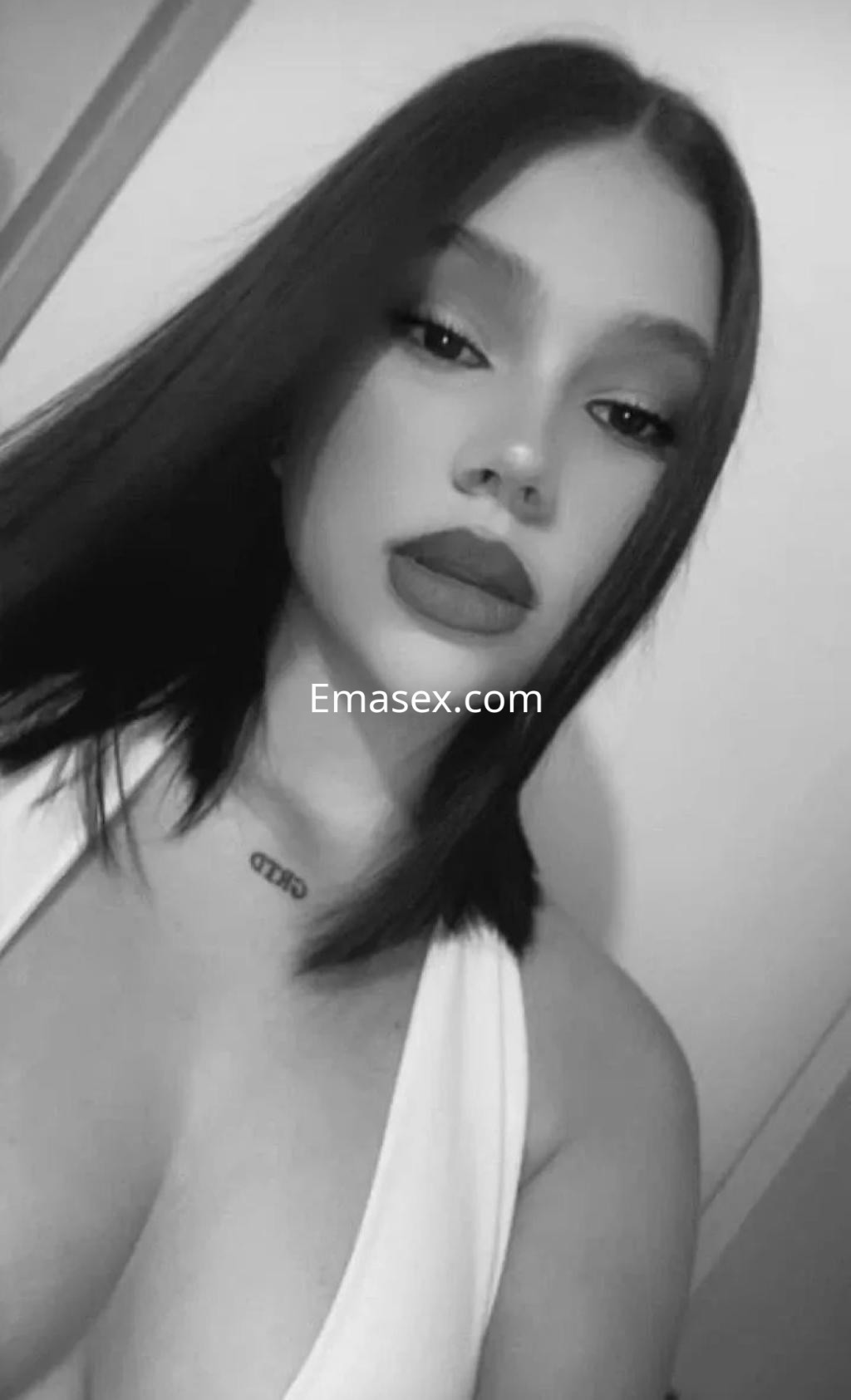 Puta con cabello lacio y oscuro, luciendo un escote en una blusa blanca, posa con labios destacados en Málaga.