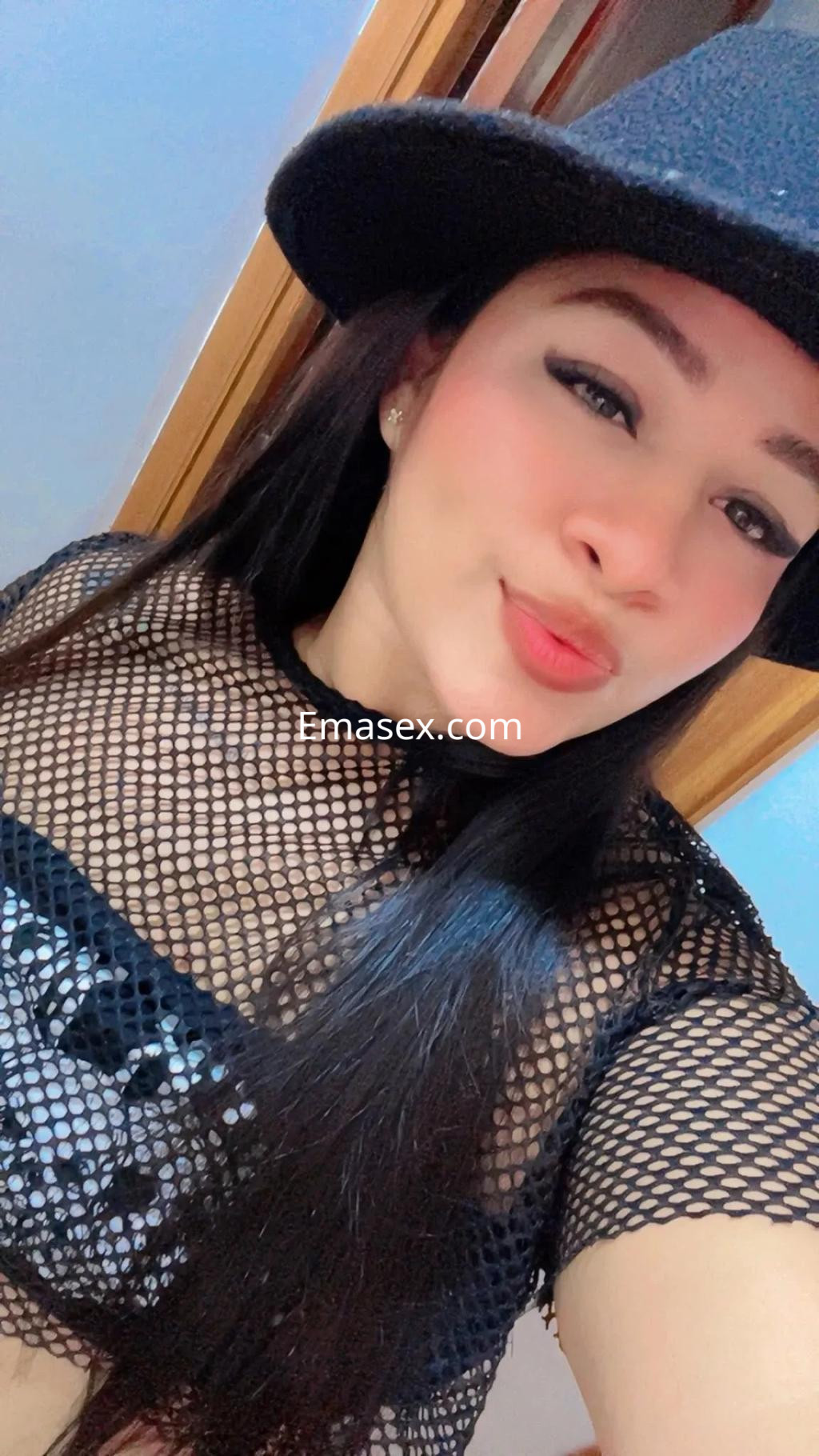 Escort en Oviedo luciendo una blusa de red negra y un sombrero, con cabello largo y lacio.