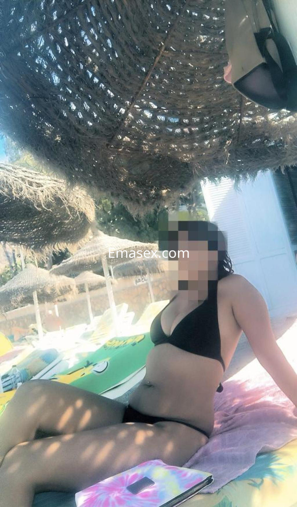 Puta en Toledo luciendo un bikini negro mientras descansa sobre una toalla en la playa, bajo una sombrilla.
