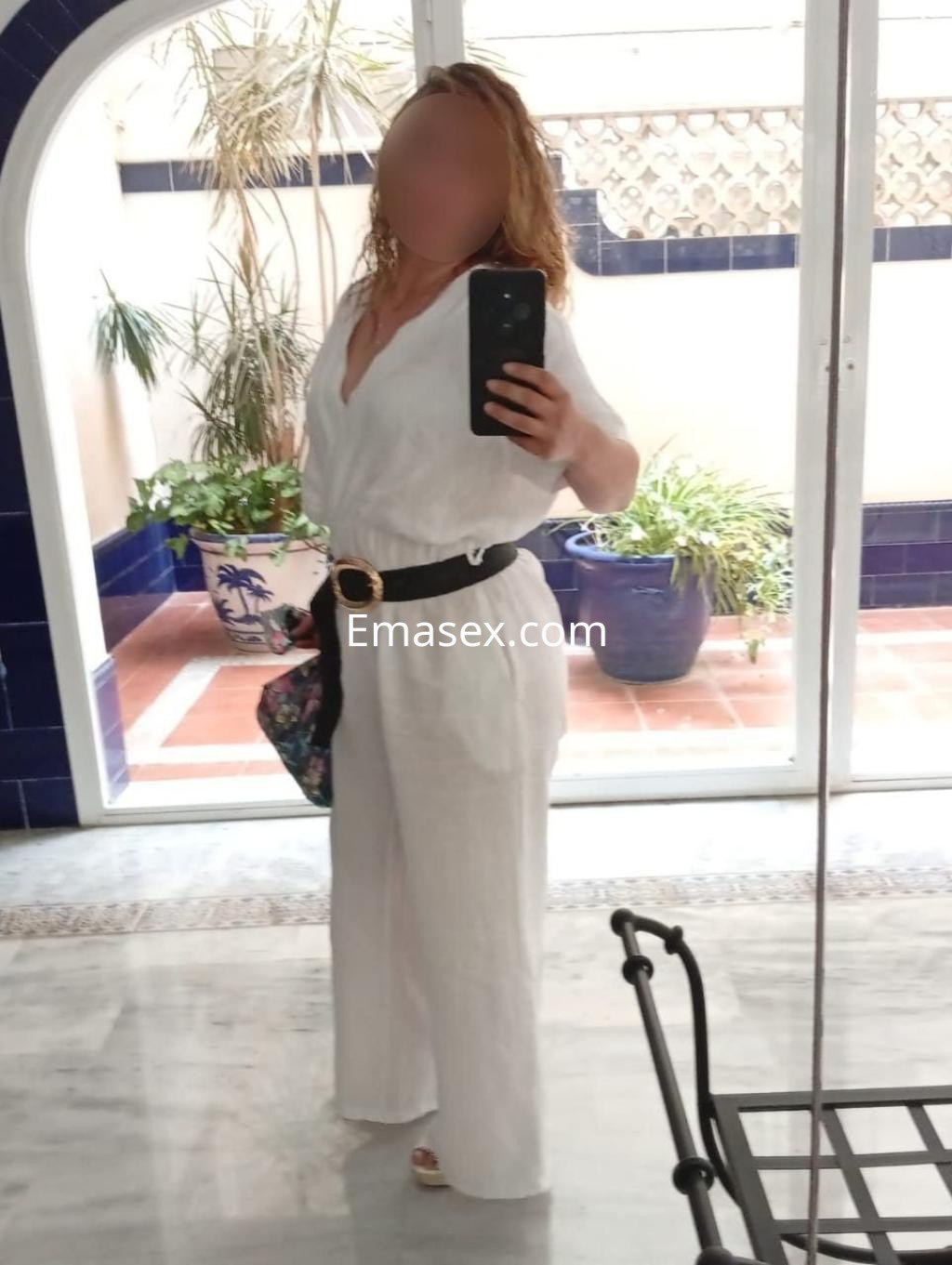 Escort en Málaga con cabello rizado y rubio, vestida con un mono blanco y cinturón negro, posando en un interior luminoso.