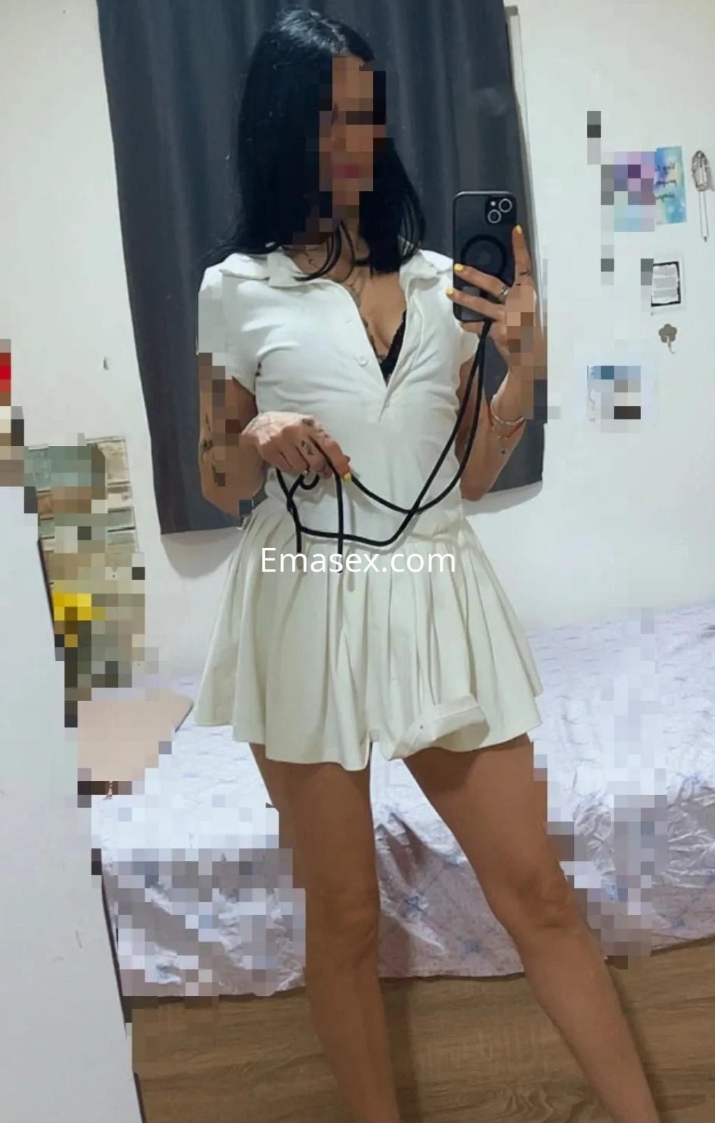 Escort en Málaga con cabello oscuro, luciendo un vestido blanco corto y posando frente a un espejo.