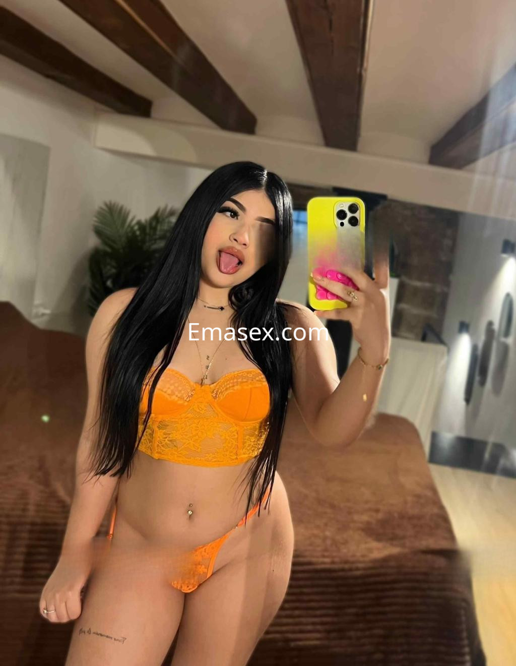 Escort en Barcelona, posando frente al espejo con lencería naranja y cabello largo y oscuro en un ambiente acogedor.