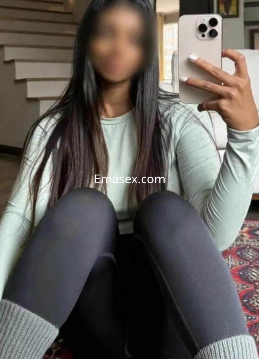 Escort en Cádiz, sentada en el suelo con leggings negros y una camiseta de manga larga verde, mostrando su teléfono.