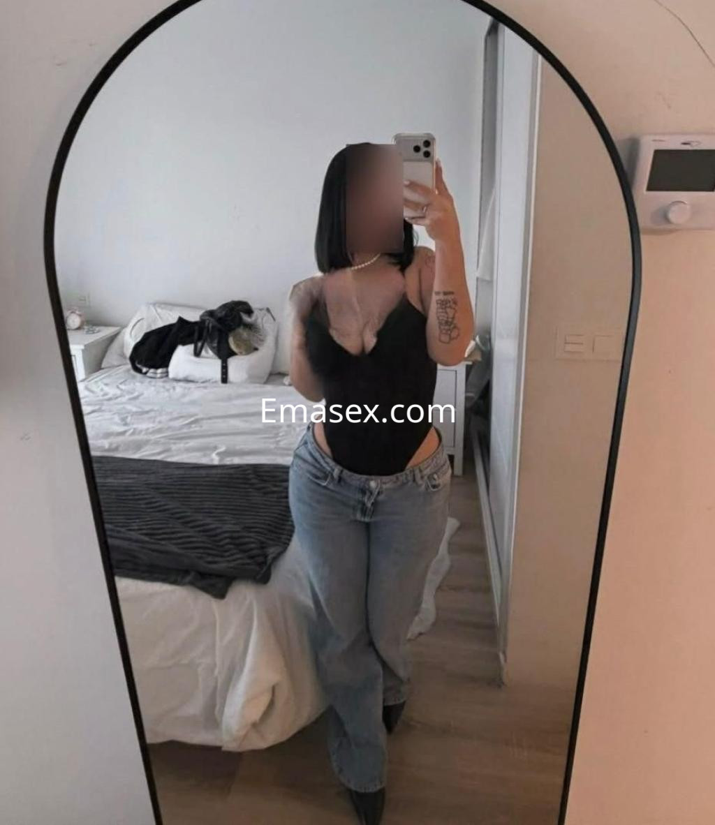 Puta en Barcelona posando frente a un espejo, con blusa negra y jeans claros en un dormitorio moderno.