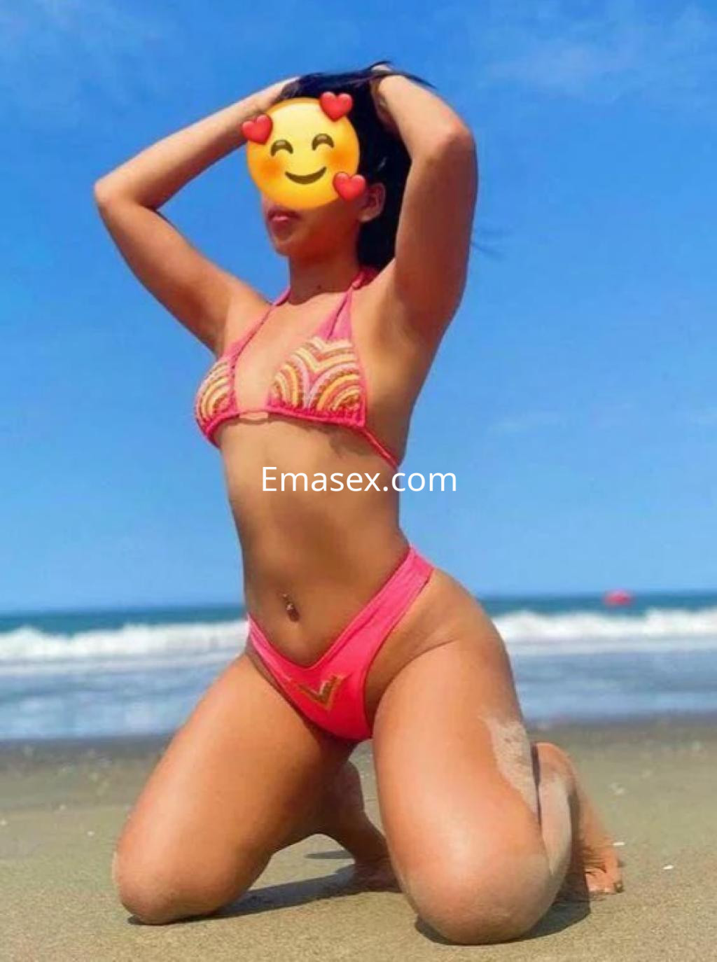 Escort en Tenerife luciendo un bikini rosa en la playa, posando de rodillas con el mar de fondo.