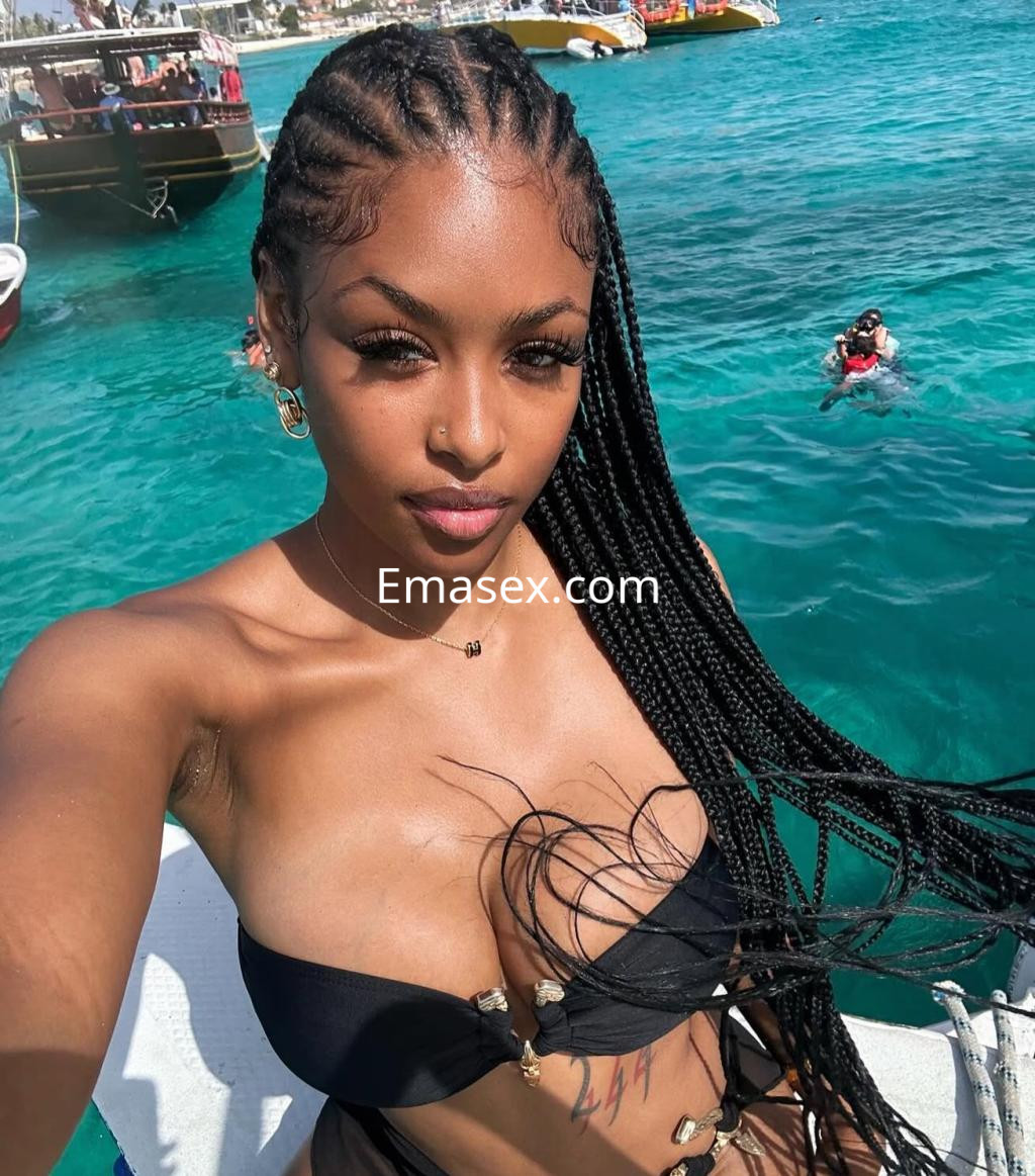 Escort en Tarragona con trenzas largas, vistiendo un bikini negro, posando frente al mar azul.