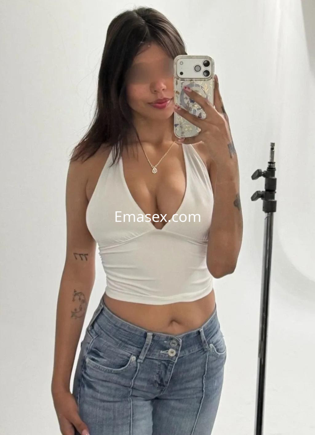La puta en Málaga posa frente al espejo con blusa blanca ajustada y jeans, mostrando confianza y sensualidad.